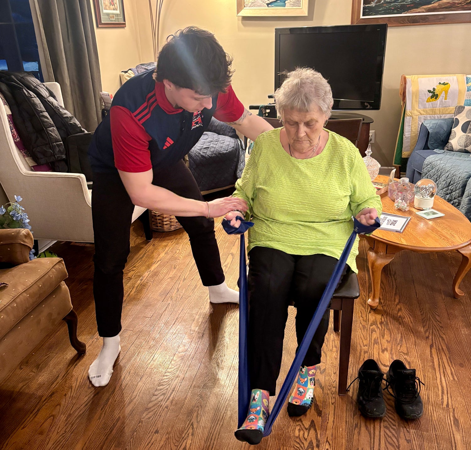 Person assisting an elderly woman with a resistance band in a living room setting.